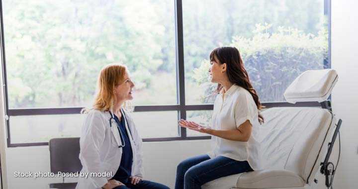 Photograph showing a female doctor in a white coat sitting and attentively listening to a female patient who is seated on an examination chair, gesturing with her hands.