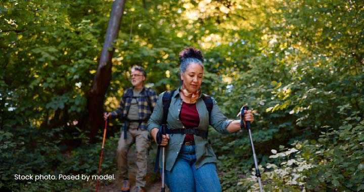 Photograph depicts two hikers trekking through a sunlit forest, with a woman leading while using trekking poles and carrying a backpack and a man following close behind.