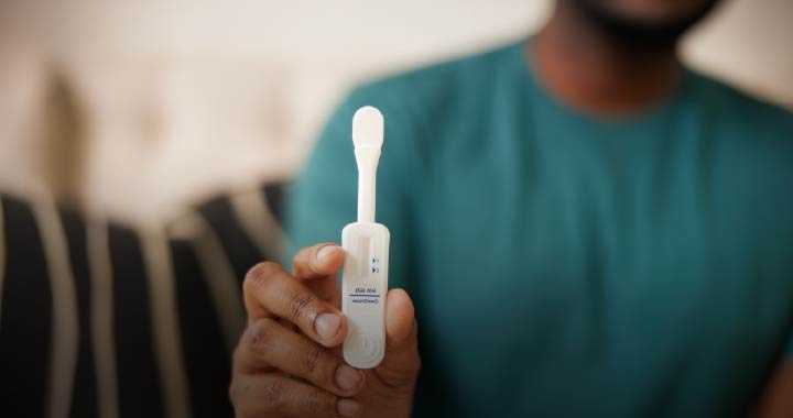 Photograph of a person holding a COVID-19 rapid antigen test device showing a negative result with one visible control line. The test device is white with blue text and symbols, and the person is wearing a green shirt with a blurred background.