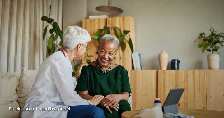 Photograph depicting a healthcare professional consulting an older woman in a warm home setting. Smiling conversation, tablet, notebook, plants, and supportive body language suggest personalized medical care or wellness counseling.